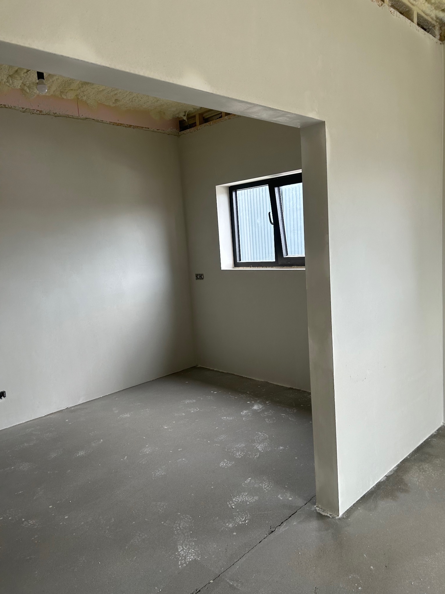 Wnętrze po nałożeniu tynków maszynowych: gładkie, jasne ściany w surowym stanie, widoczne ślady stóp na szarej posadzce, okno z czarną ramą i pianka montażowa pod sufitem.