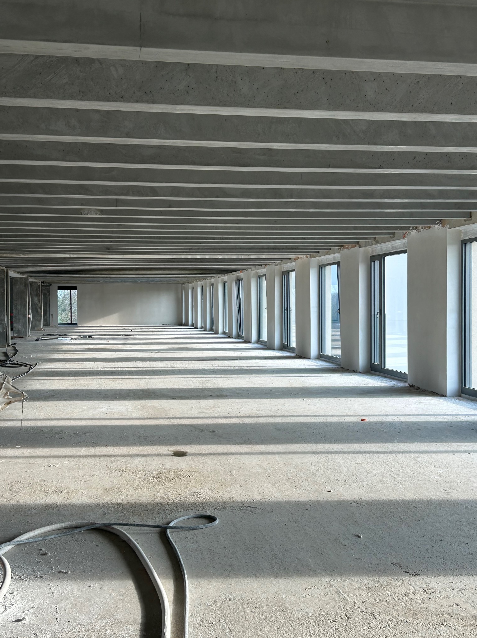 Wnętrze surowego, betonowego budynku w trakcie prac wykończeniowych, widoczne betonowe belki stropowe, okna w szpalerze i szary wąż na posadzce.