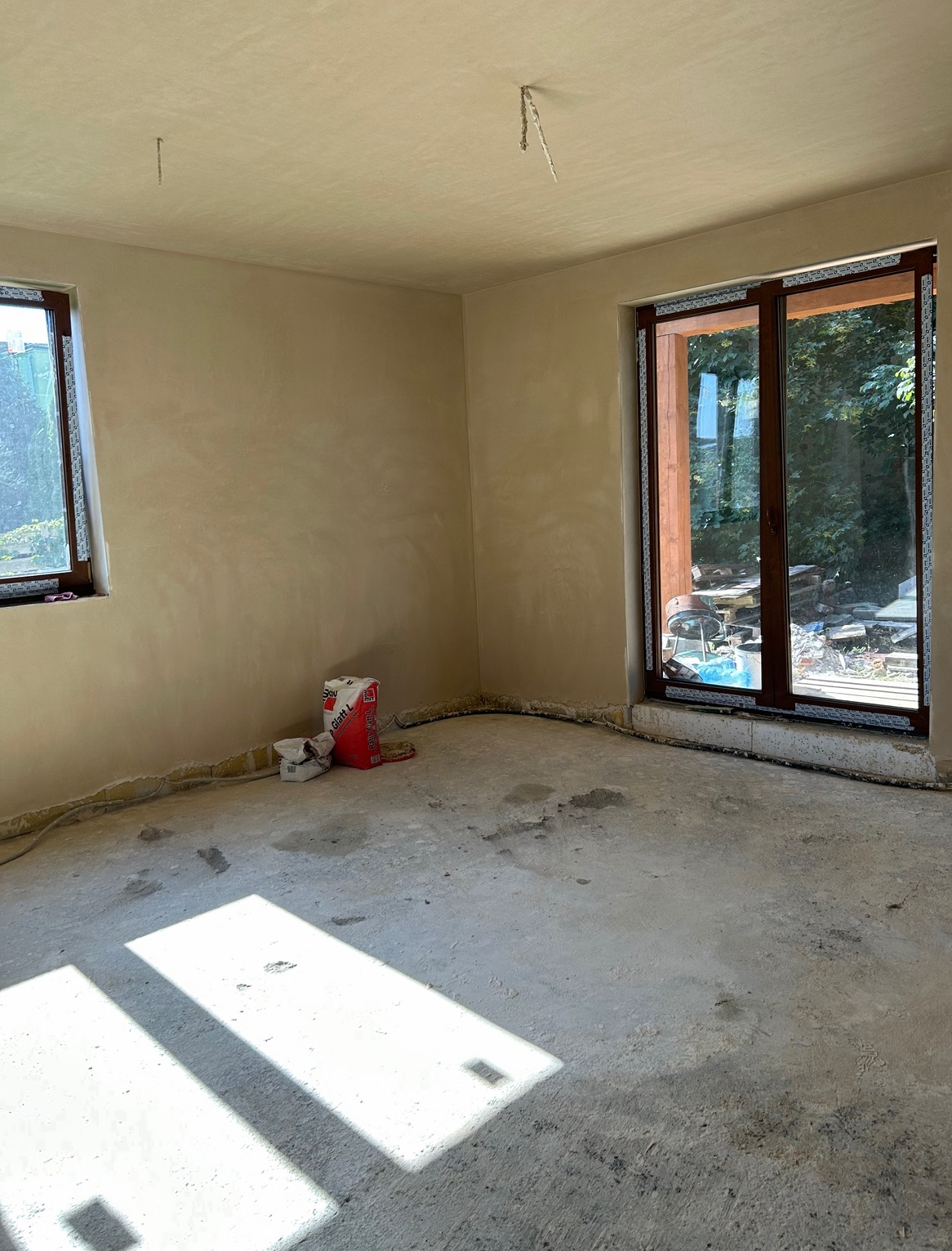 Wnętrze pokoju po nałożeniu gładzi, widoczne okna, drzwi balkonowe, worki z materiałem i przewody elektryczne przy podłodze.