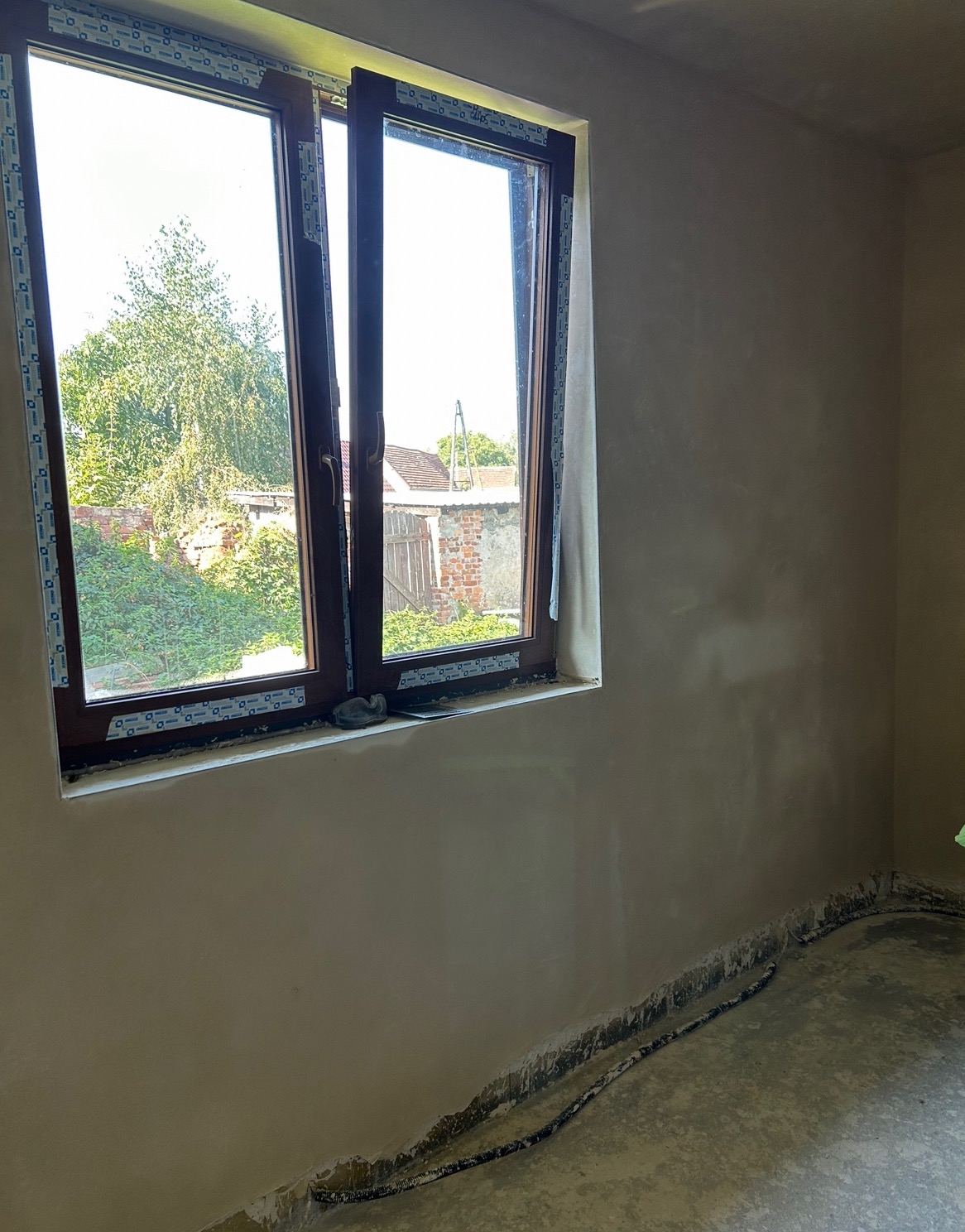 Świeżo nałożony tynk na ścianie wewnętrznej, widoczne okno z widokiem na ogród, szpachelka leżąca na parapecie.