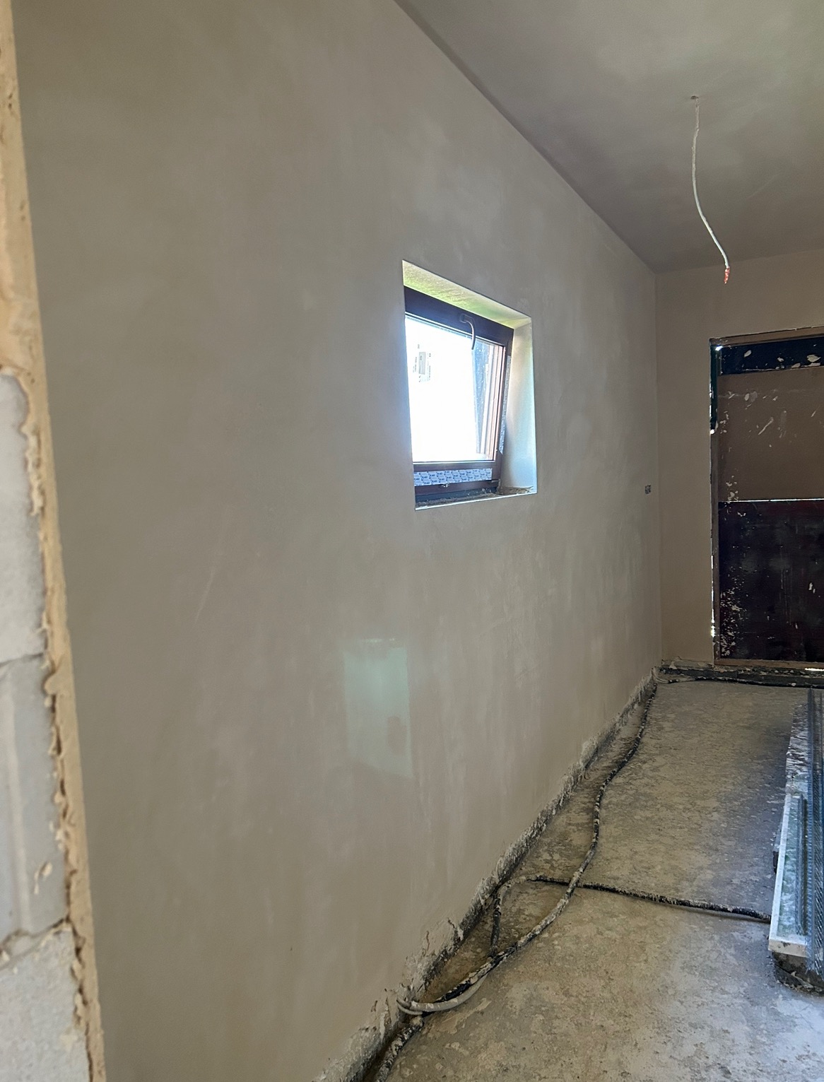 Wnętrze po nałożeniu gładzi gipsowej, widoczne okno, kable przy podłodze i surowy stan wykończenia.