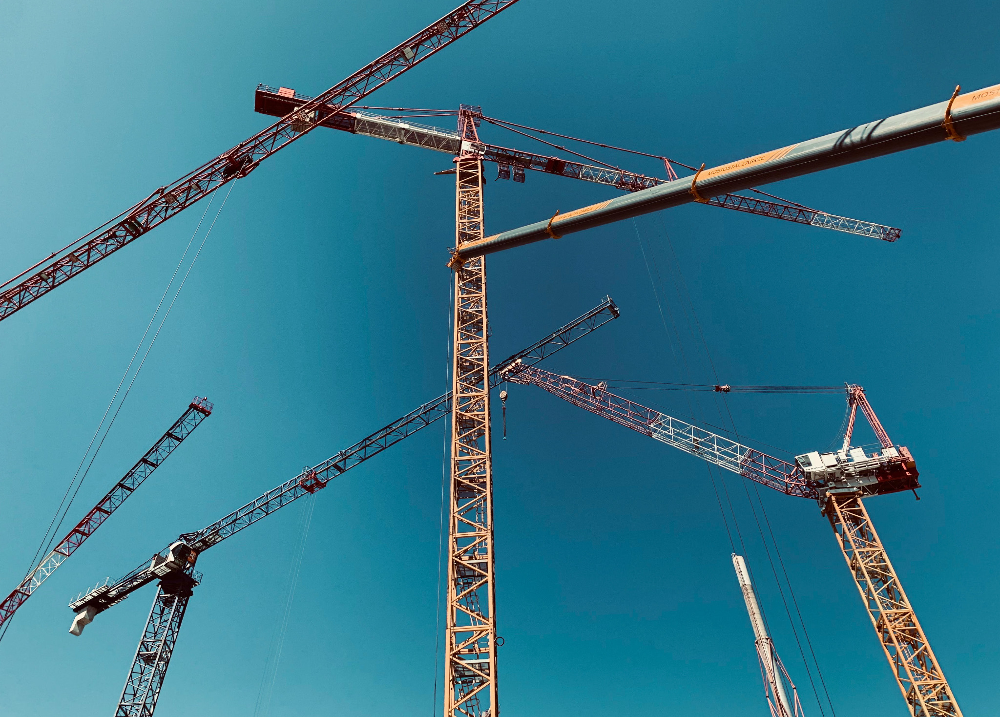 Kilka żurawi budowlanych o różnych kolorach konstrukcji (czerwony, żółty, szary) na tle intensywnego błękitu nieba, ujęcie z dołu podkreśla ich wysokość i złożoność konstrukcji.