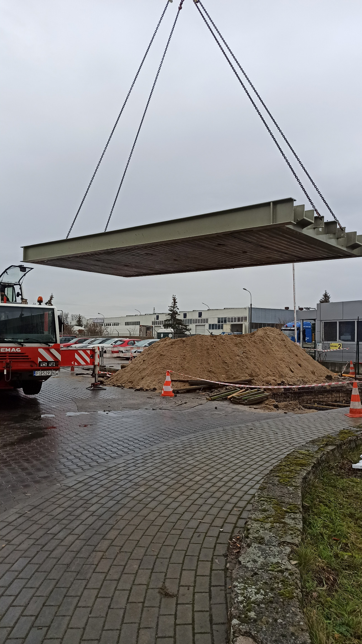 Montaż prefabrykowanego elementu konstrukcyjnego dźwigiem na budowie w Stargardzie, widoczny stos piasku i zaparkowany pojazd budowlany.