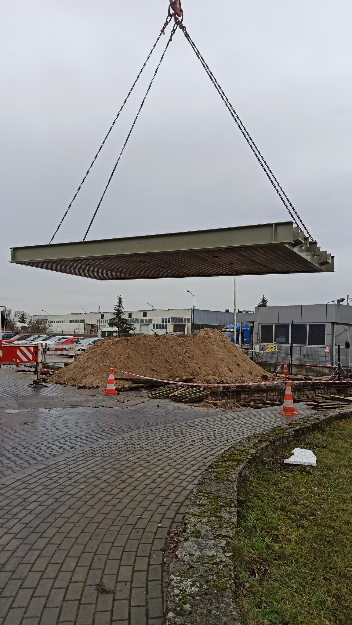 Montaż konstrukcji stalowej za pomocą dźwigu nad stertą piasku na placu budowy, widoczne elementy zabezpieczeń i budynki w tle.