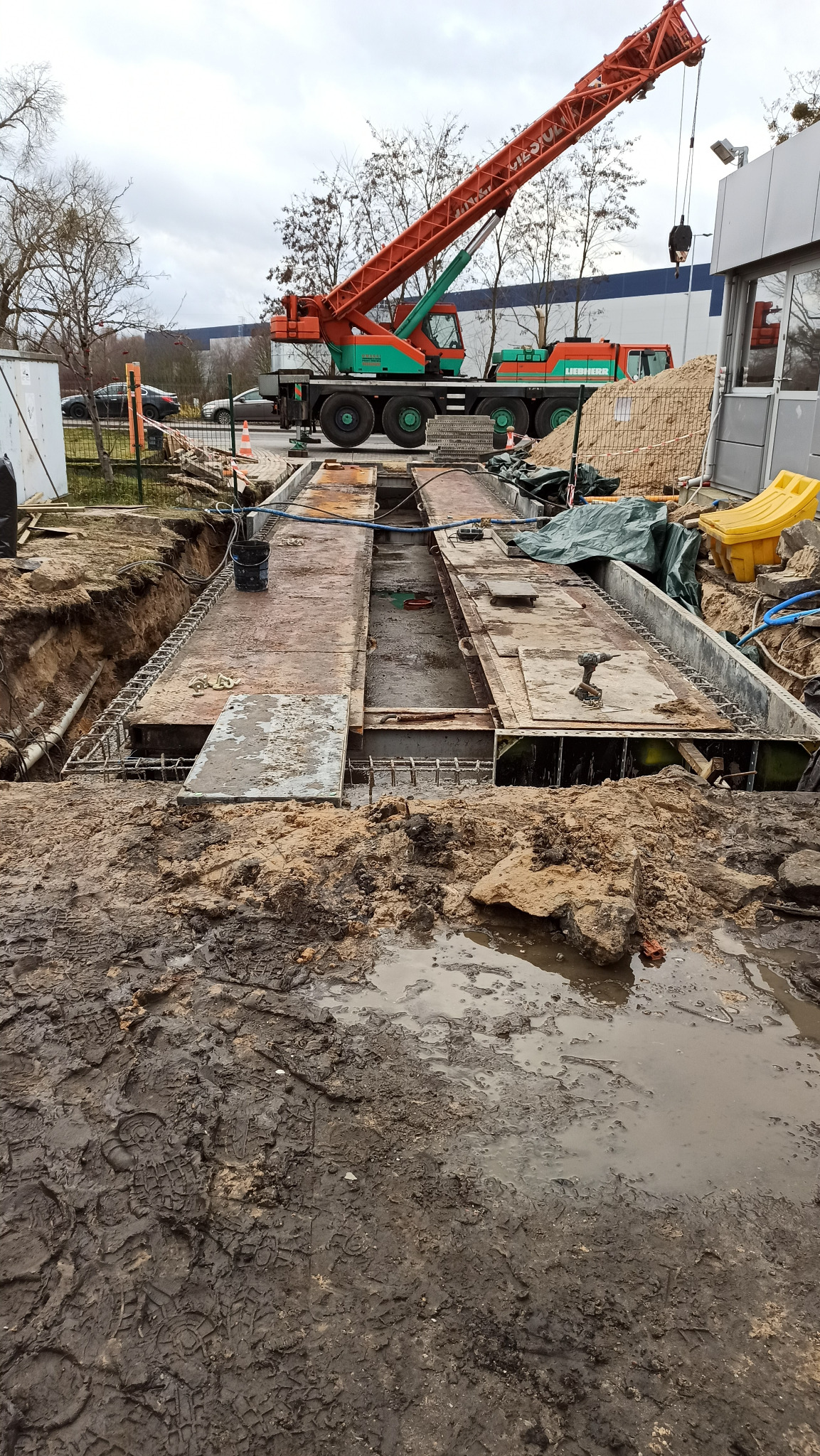 Fundamenty pod budynek w trakcie wylewania betonu, widoczny dźwig Liebherr w tle, błotniste podłoże z odciskami butów.