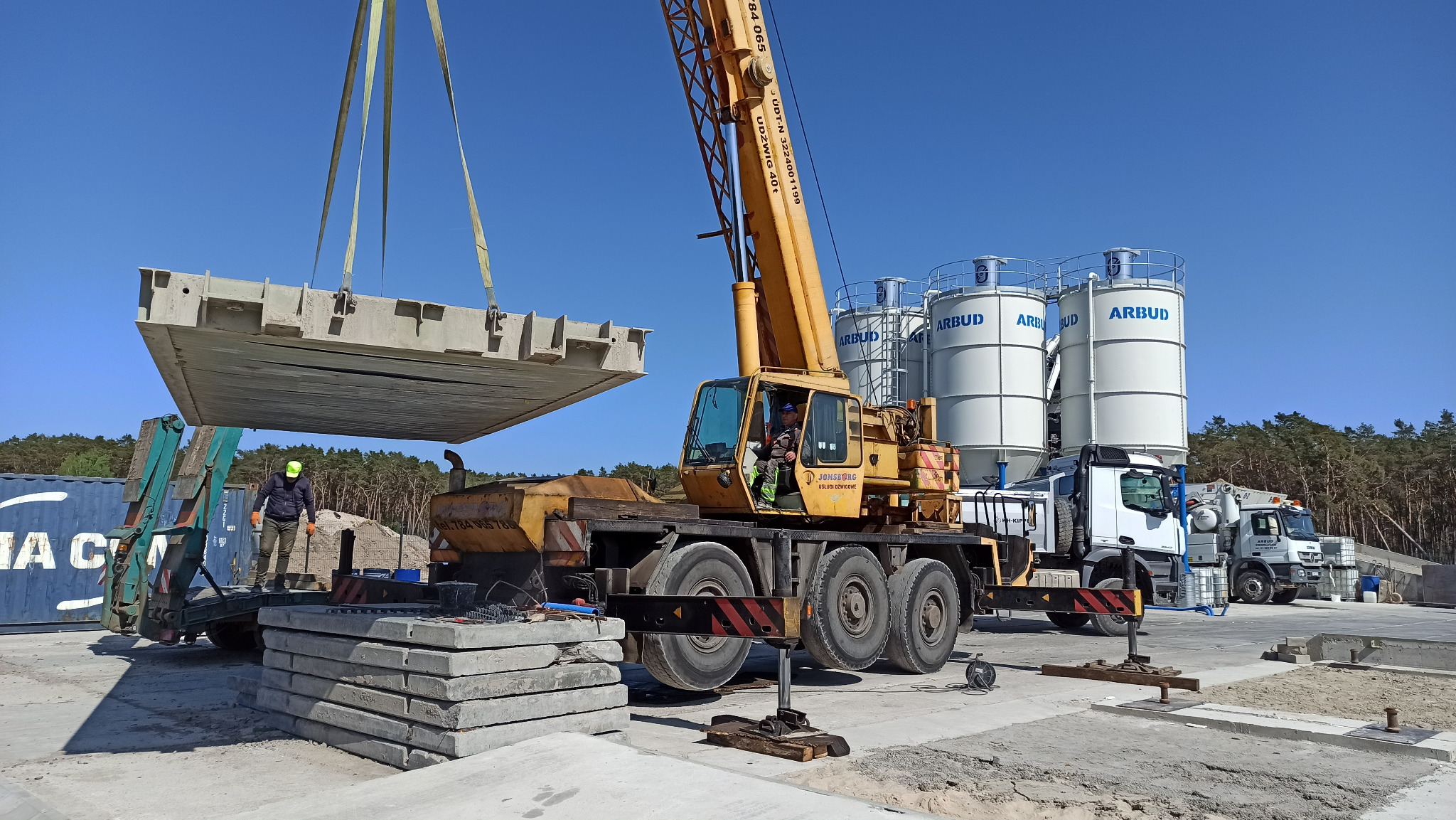 Żółty dźwig podnosi betonowy prefabrykat na placu budowy, widoczne silosy z napisem ARBUD, pracownik w zielonej czapce i sterta płyt betonowych.
