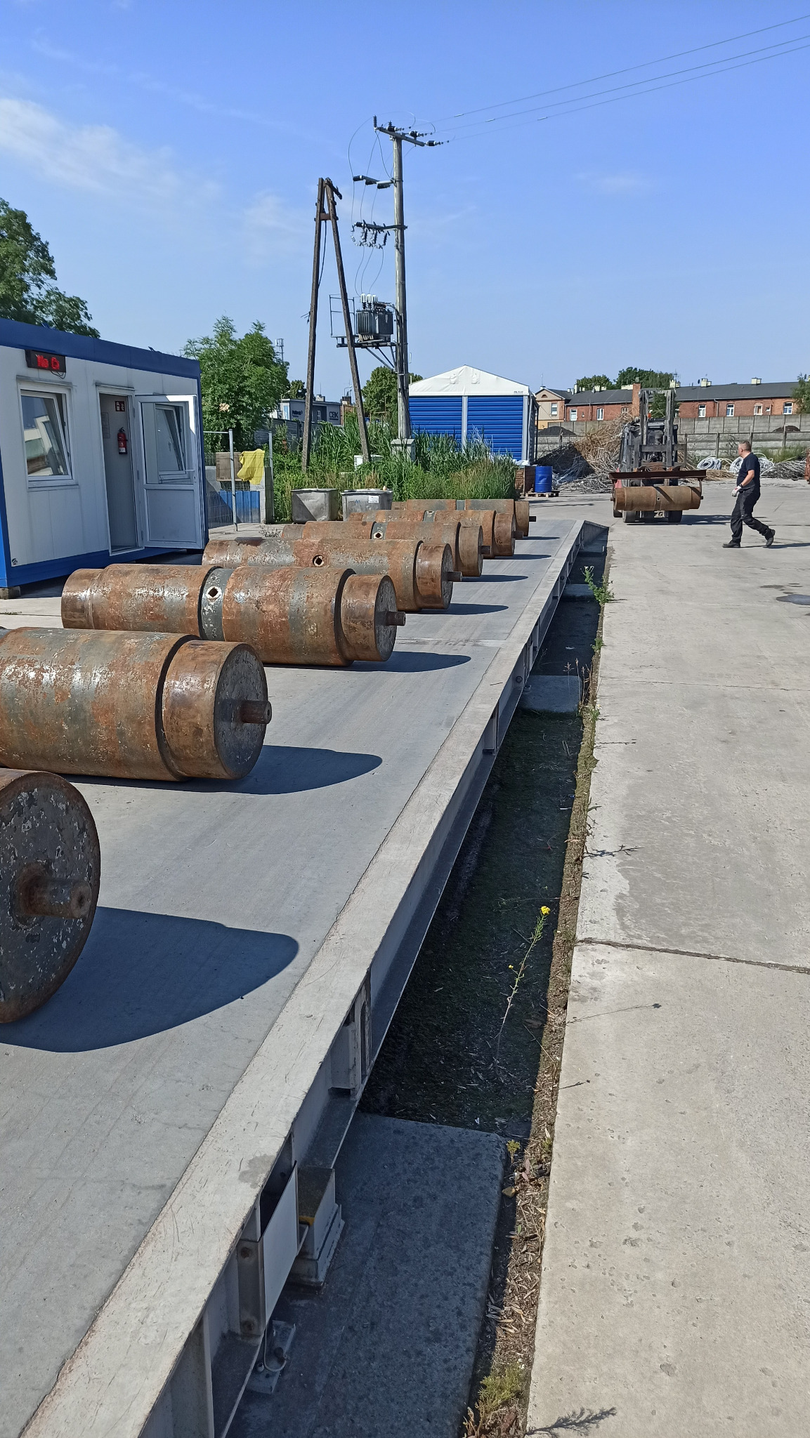 Rząd zardzewiałych, metalowych cylindrów na betonowej platformie, z widocznym w tle wózkiem widłowym i przechodzącym mężczyzną, w tle budynki i słupy energetyczne.