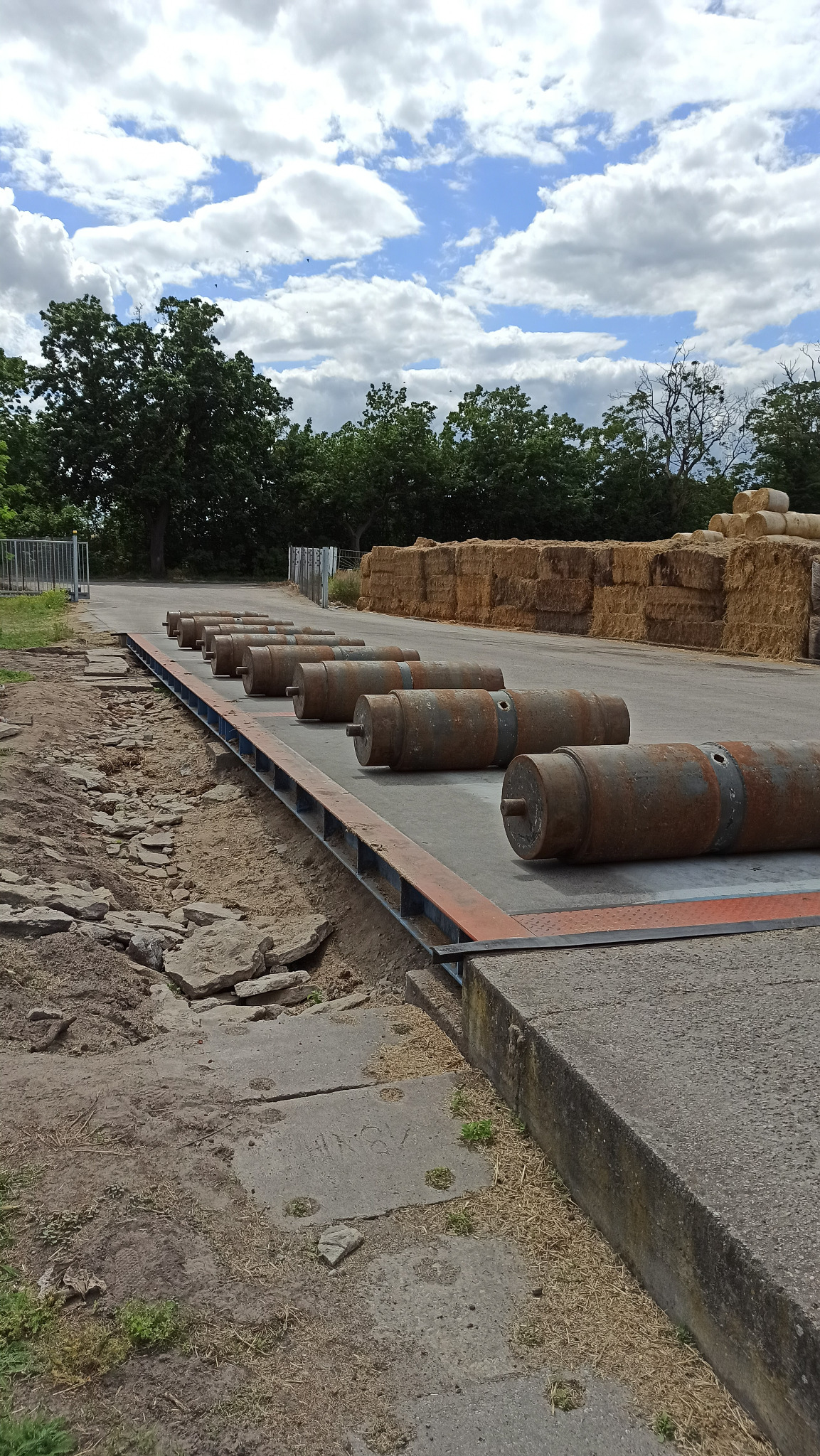 Rząd zardzewiałych, cylindrycznych obciążników na betonowej wadze najazdowej, z belami słomy w tle i fragmentem nieba z chmurami.