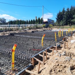 HOVERLA LTD - Zbrojenie fundamentu z żółtymi rurami osłonowymi wystającymi pionowo, widok z góry na siatkę zbrojeniową i drewniane szalunki, w tle budynek i drzewa.