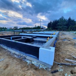 HOVERLA LTD - Zabezpieczone fundamenty domu w trakcie budowy, z czarną hydroizolacją i białym styropianem, widoczne pręty zbrojeniowe i żółte znaczniki, tło pochmurne niebo.