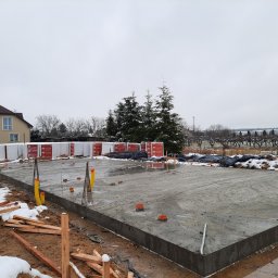 HOVERLA LTD - Wylana płyta fundamentowa z wystającymi rurami i prętami zbrojeniowymi, w tle palety z materiałami budowlanymi i częściowo ośnieżony krajobraz.