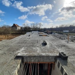 HOVERLA LTD - Wylana płyta fundamentowa z widocznymi otworami i wystającymi prętami zbrojeniowymi, w tle zabudowa jednorodzinna i błękitne niebo z chmurami.