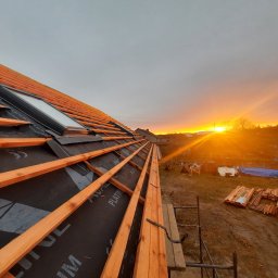 HOVERLA LTD - Konstrukcja dachu z oknem dachowym, pokryta membraną, przygotowana pod montaż dachówki, widok na zachodzące słońce.