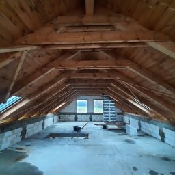 HOVERLA LTD - Surowe poddasze z drewnianą konstrukcją dachu, oknami dachowymi i murem z bloczków betonowych w trakcie prac, widoczne ślady wilgoci na podłodze i drabina oparta o ścianę.