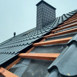 HOVERLA LTD - Czarny dach z blachy trapezowej w trakcie montażu, widoczne drewniane łaty i komin obłożony łuską, pochmurne niebo w tle.