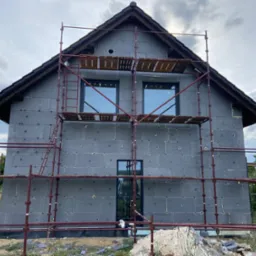 Dwukondygnacyjny dom w trakcie ocieplania styropianem, widoczne rusztowania, okna i dach pokryty dachówką.