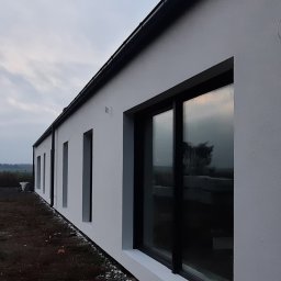 HOVERLA LTD - Świeżo otynkowany, parterowy budynek jednorodzinny z czarną stolarką okienną i orynnowaniem, widoczny z niskiej perspektywy, z niezagospodarowanym terenem wokół.