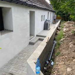 HOVERLA LTD - Biała ściana budynku z szarymi elementami chodnika i schodów, widoczna instalacja klimatyzacji i fragment terenu z rurą, w tle mężczyzna z narzędziem.