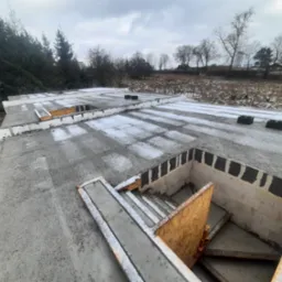 Wylany betonowy strop z widocznym otworem na schody, zabezpieczonym płytą OSB, lekko przyprószony śniegiem, w tle drzewa i pochmurne niebo.