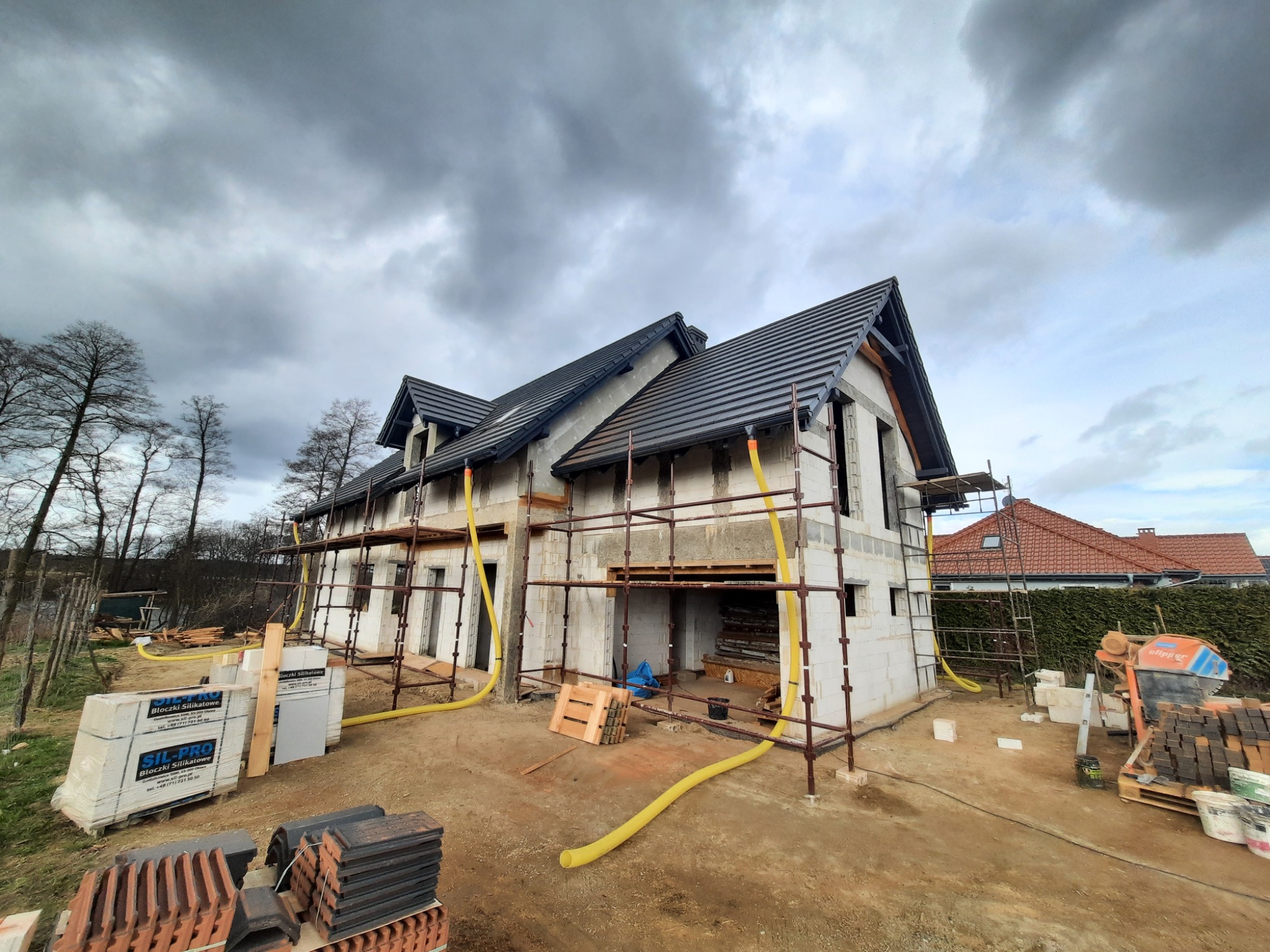 Surowy dom murowany w trakcie budowy, otoczony rusztowaniami i materiałami budowlanymi, takimi jak bloczki silikatowe i dachówki, na tle pochmurnego nieba.