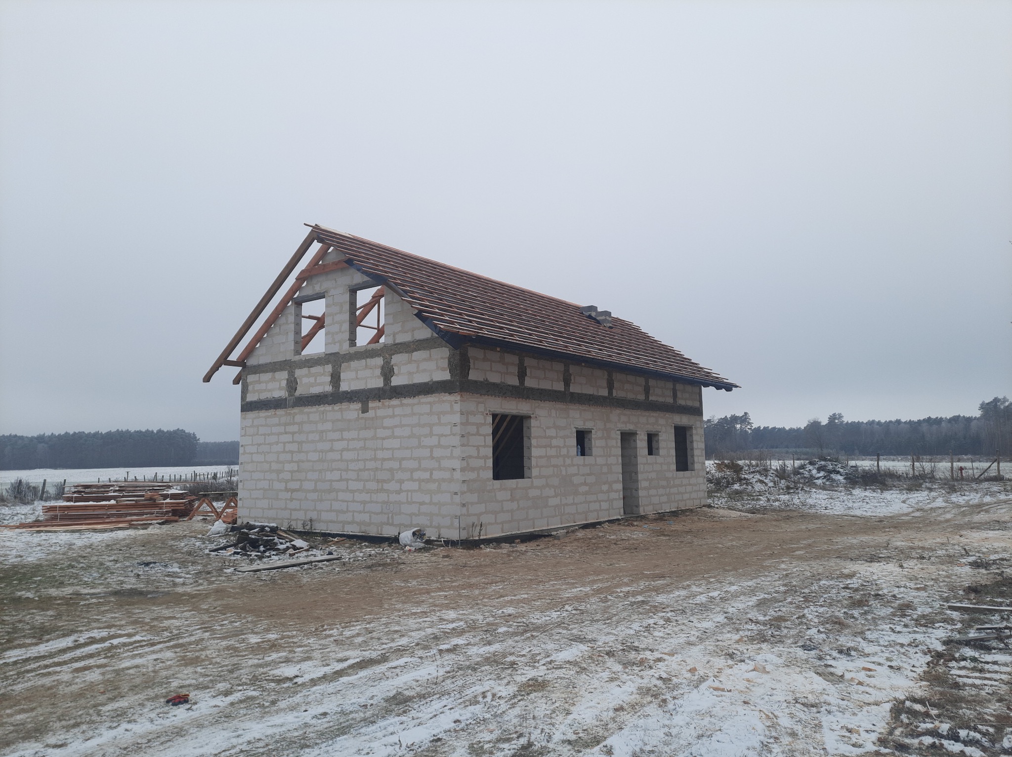 Surowy stan dwukondygnacyjnego budynku z białych bloczków z częściowo ułożoną czerwoną dachówką na tle zimowego krajobrazu, widoczne przygotowane deski i drewniana konstrukcja dachu.