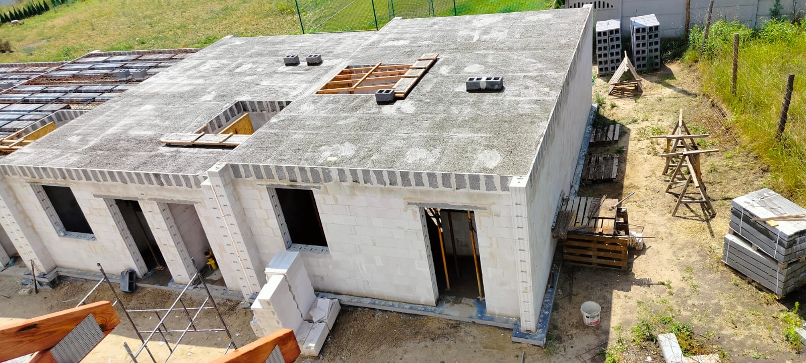 Surowy stan parterowego budynku z białych bloczków, widok z góry ukazuje płaski dach z otworami na okna dachowe, częściowo zbrojony strop i elementy drewniane konstrukcji więźby dachowej,...