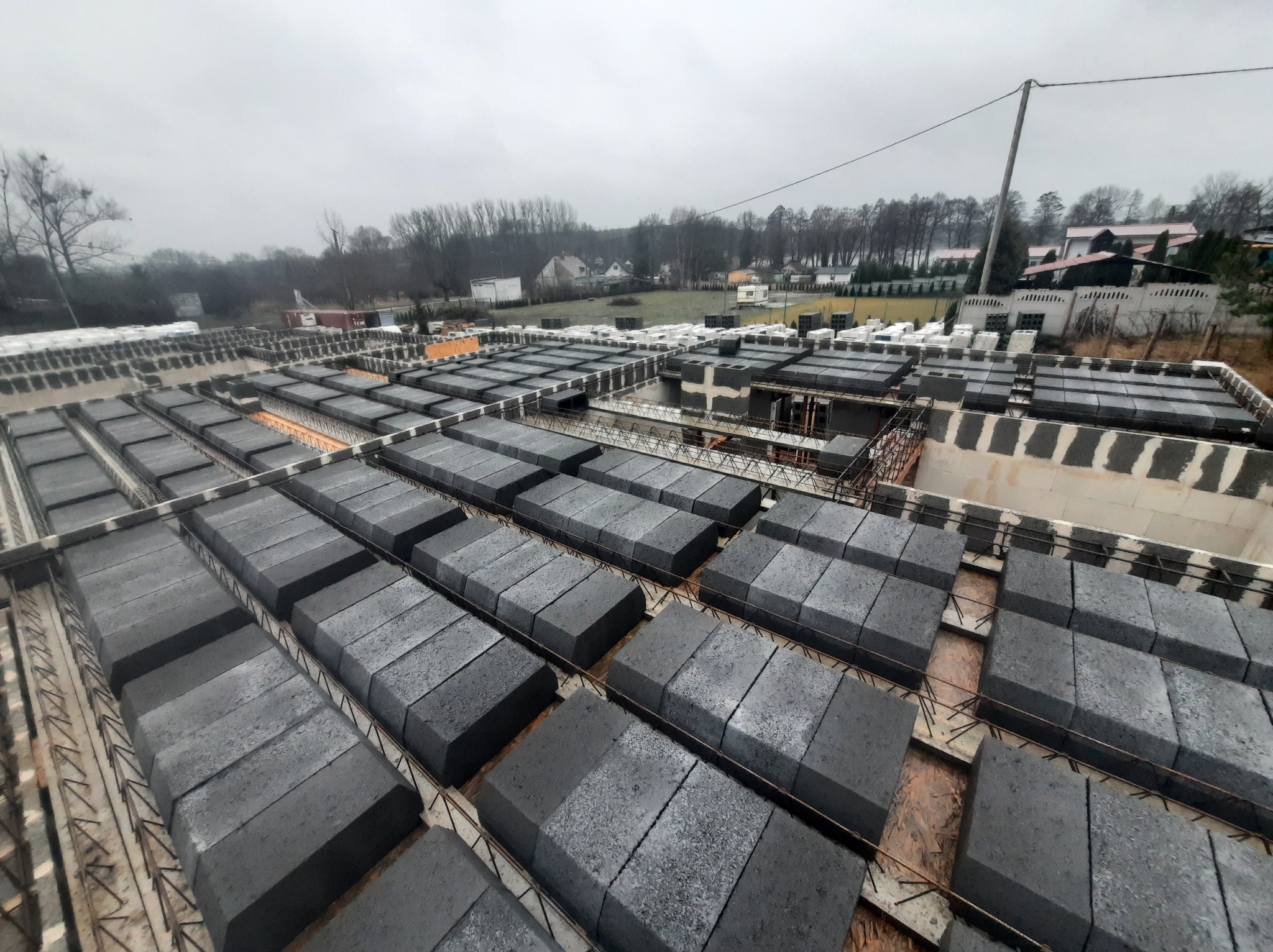 Widok z góry na budowę domu z szarej cegły, z widocznymi elementami konstrukcyjnymi i zbrojeniami, w tle zabudowa jednorodzinna i pochmurne niebo.