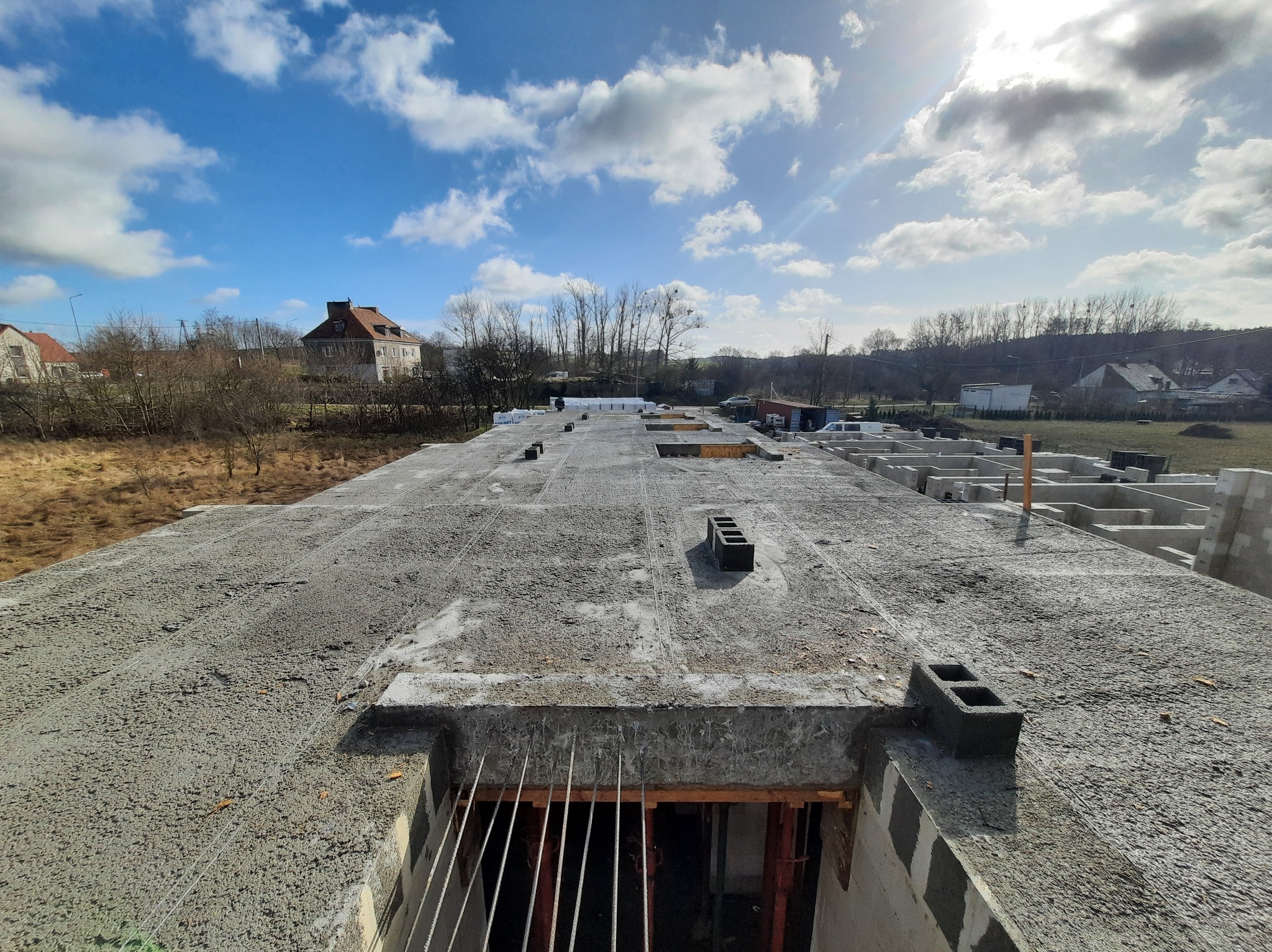 Wylana płyta fundamentowa z widocznymi otworami i wystającymi prętami zbrojeniowymi, w tle zabudowa jednorodzinna i błękitne niebo z chmurami.