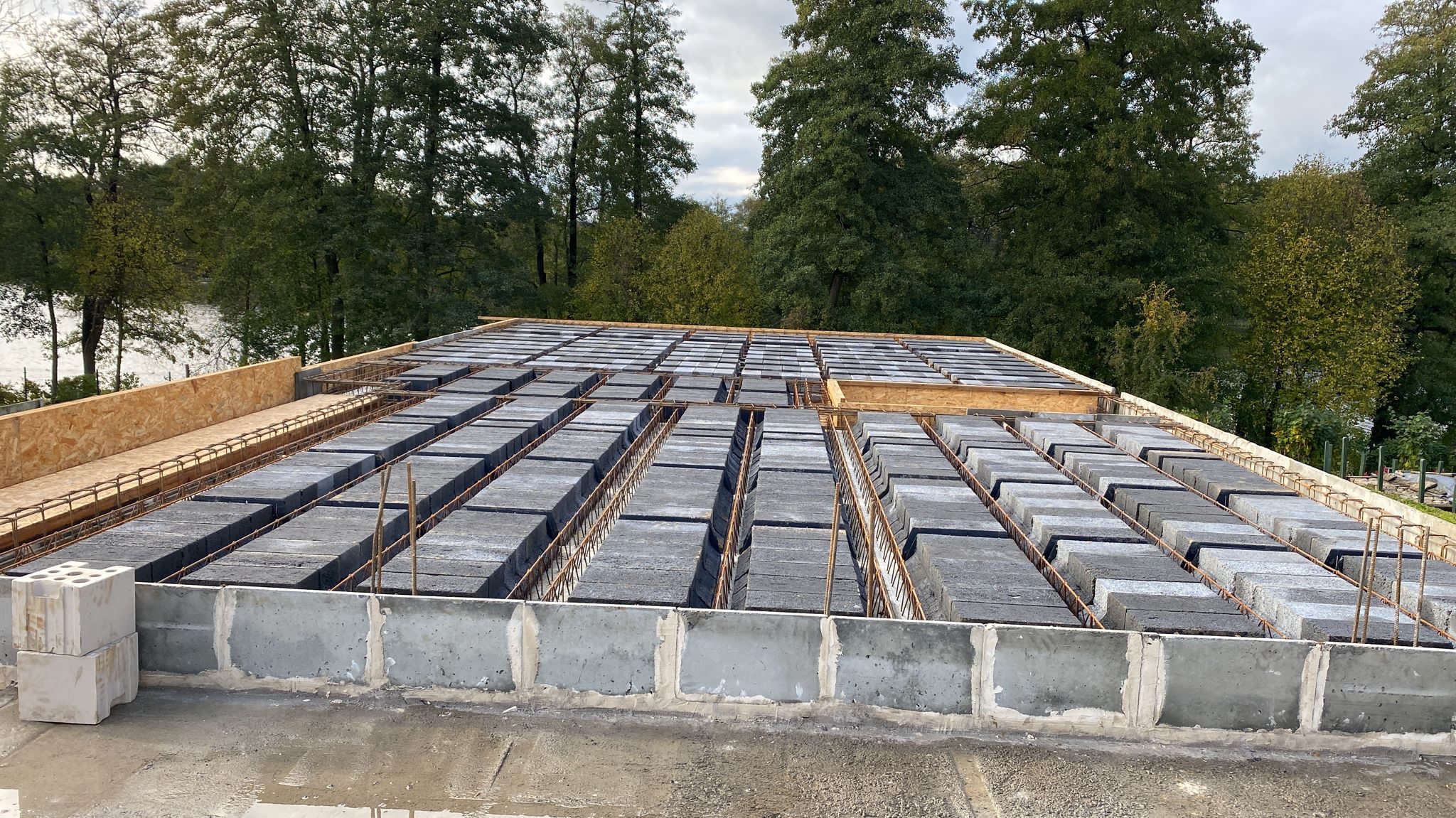 Wykonywanie stropu Teriva na budowie domu jednorodzinnego, widoczne bloczki betonowe ułożone między żebrami oraz zbrojenie, w tle drzewa i jezioro.