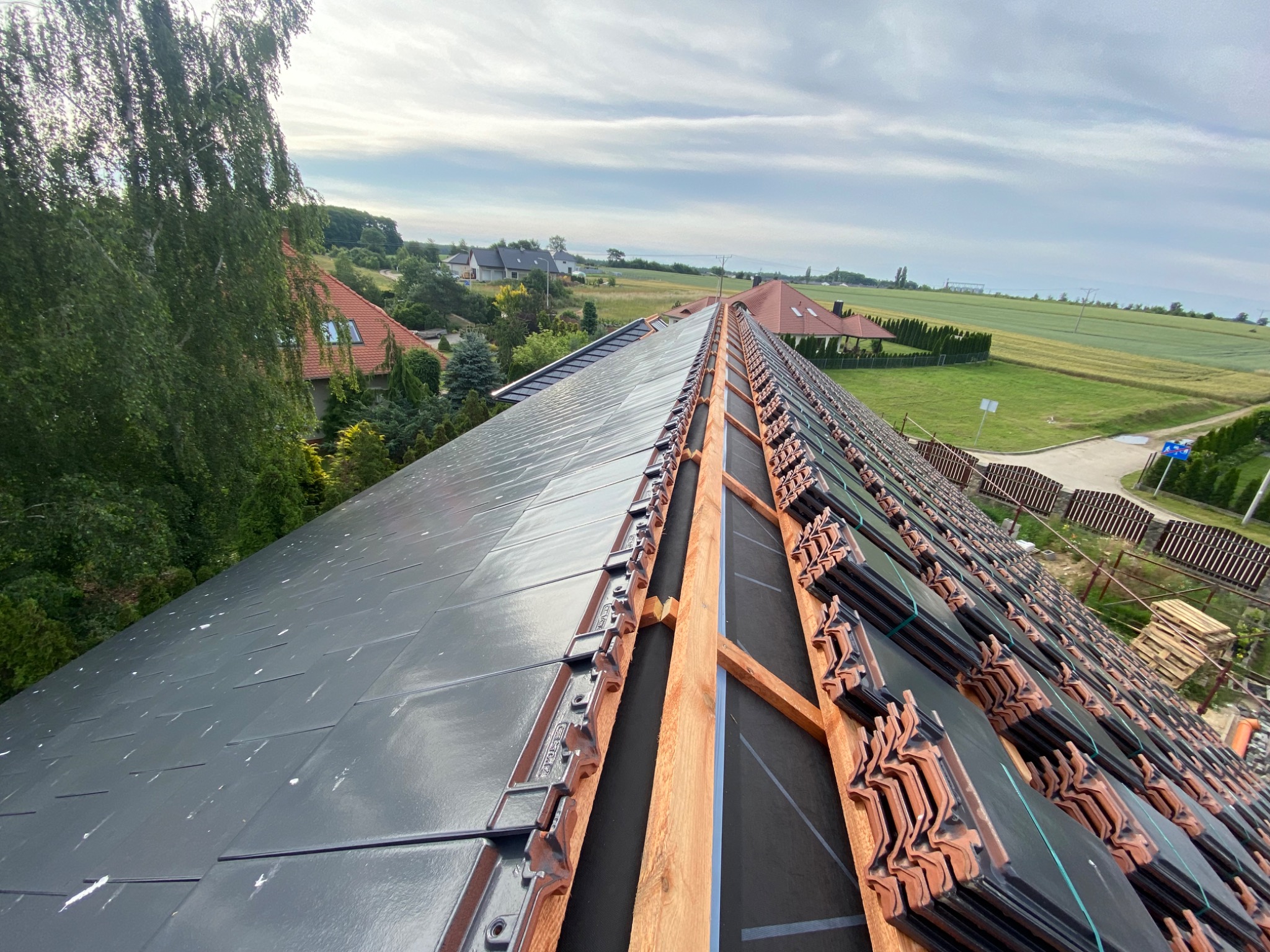 Montaż dachówki ceramicznej na dachu domu jednorodzinnego z widokiem na okolicę, widoczne drewniane łaty i membrany dachowe.