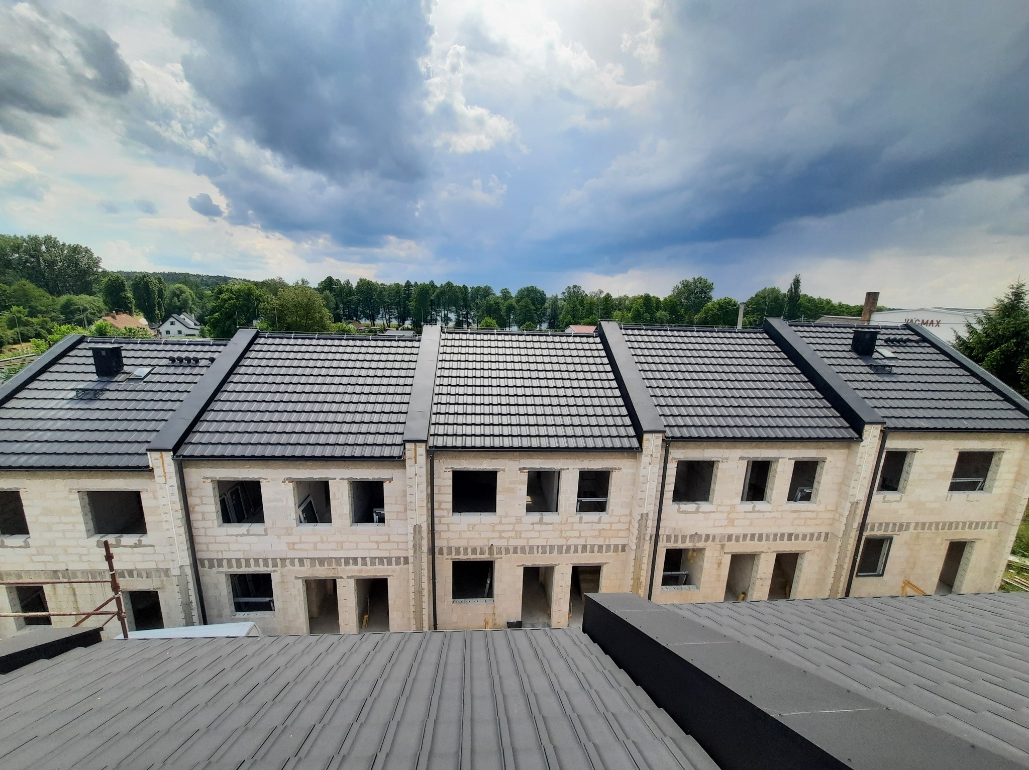 Wysoki kąt widzenia na szereg domów w stanie surowym zamkniętym, z szarą dachówką i nieotynkowanymi ścianami z betonu komórkowego, na tle pochmurnego nieba.