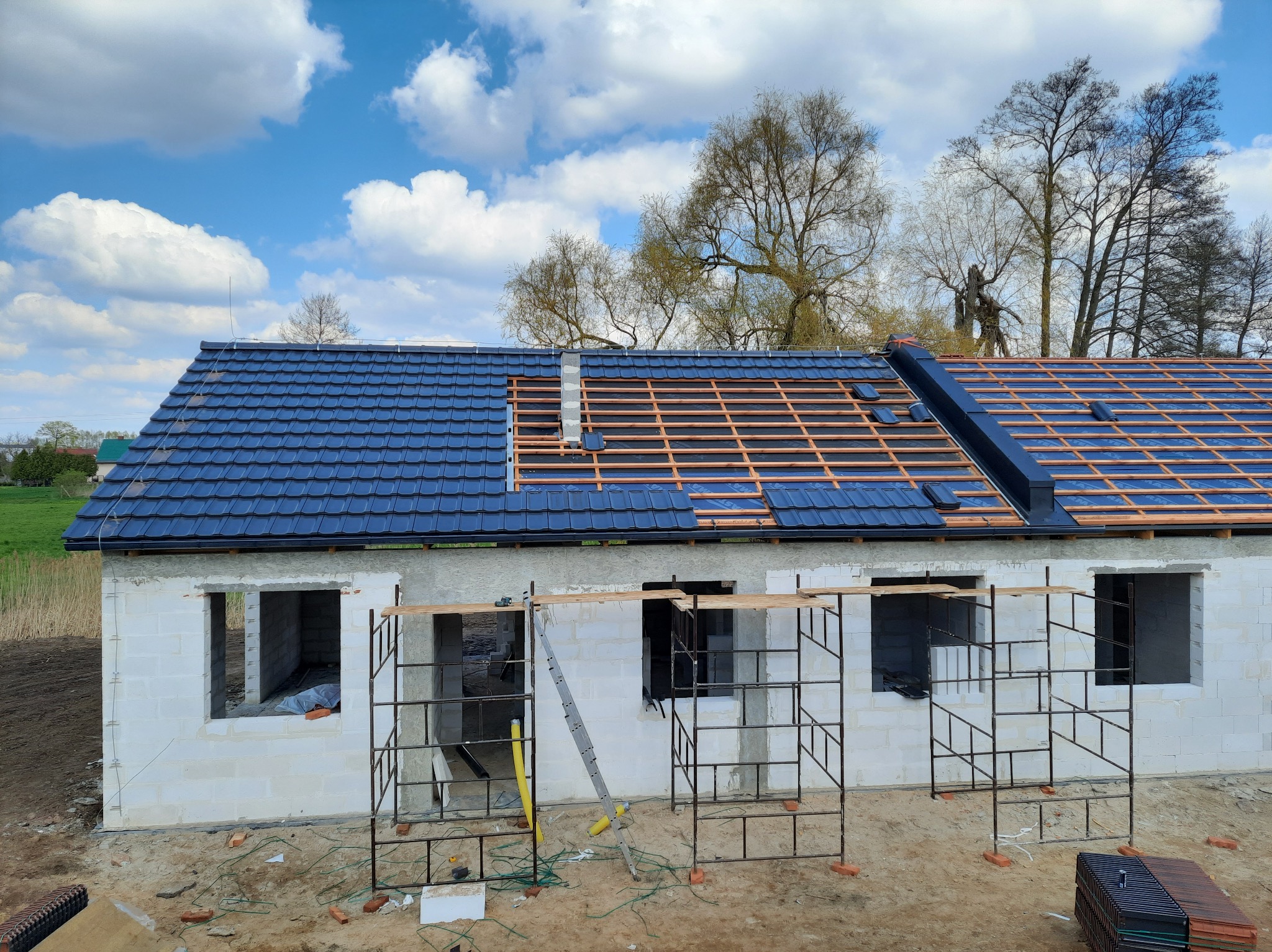 Niedokończony budynek z białych bloczków, z częściowo ułożonym, ciemnoniebieskim dachem z dachówki, widoczne drewniane łaty, rusztowania i drabina oparta o ścianę, pod błękitnym niebem z chmurami.