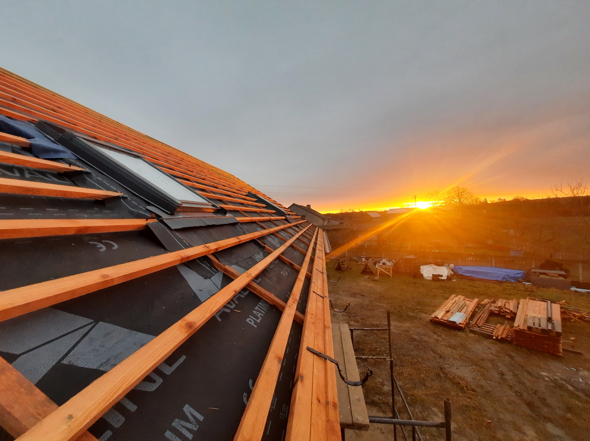 Konstrukcja dachu z oknem dachowym, pokryta membraną, przygotowana pod montaż dachówki, widok na zachodzące słońce.