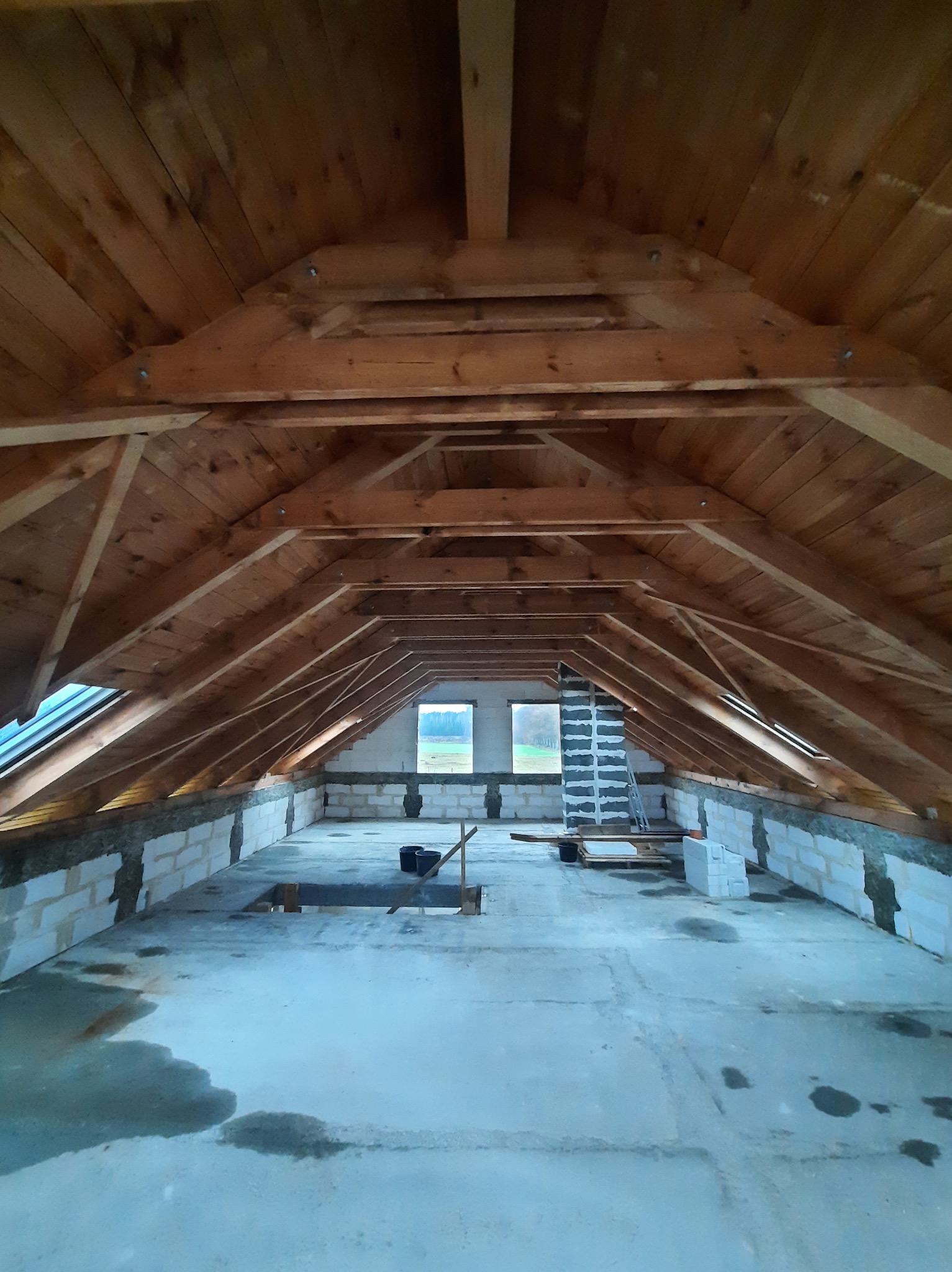 Surowe poddasze z drewnianą konstrukcją dachu, oknami dachowymi i murem z bloczków betonowych w trakcie prac, widoczne ślady wilgoci na podłodze i drabina oparta o ścianę.