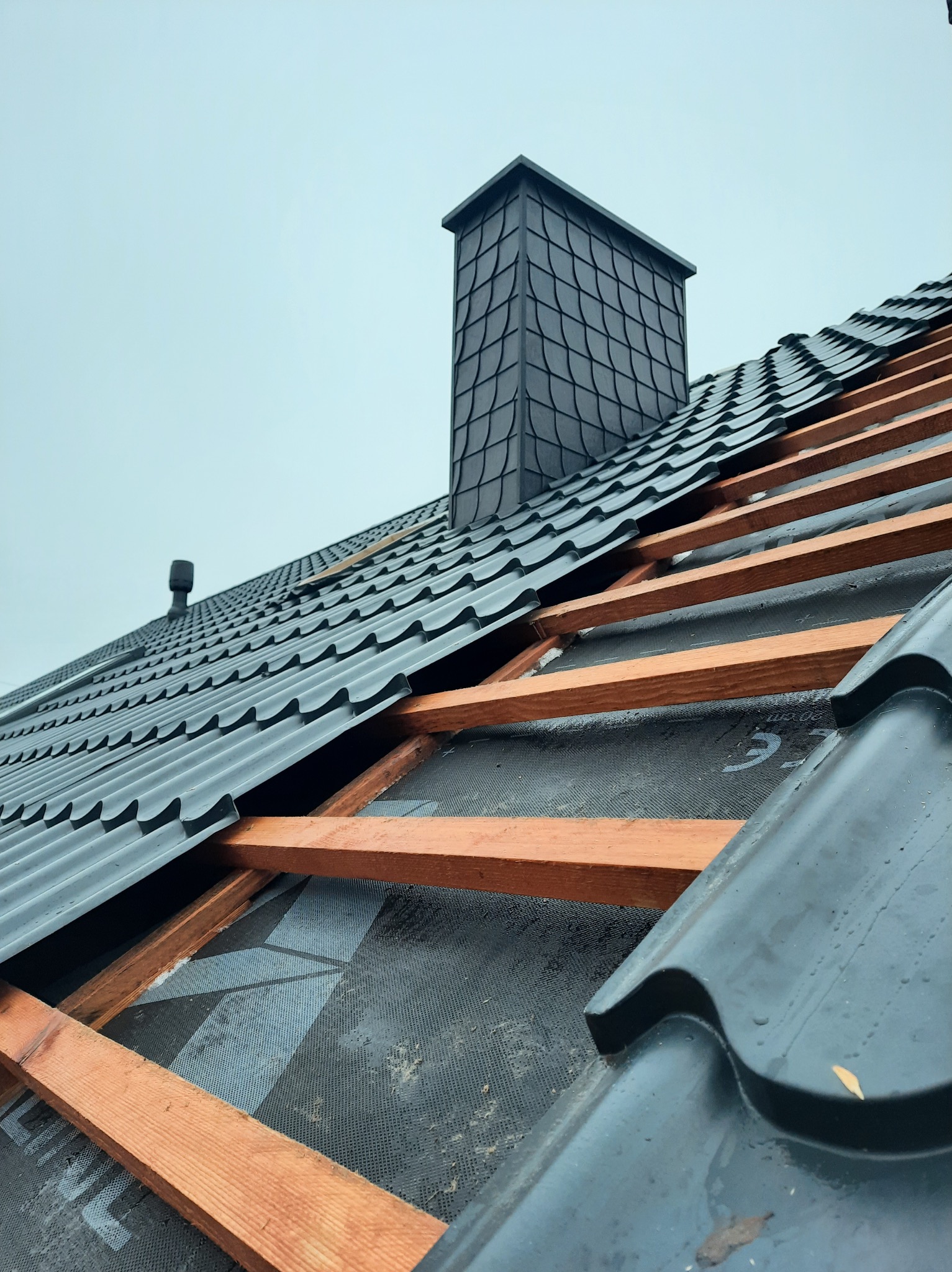 Czarny dach z blachy trapezowej w trakcie montażu, widoczne drewniane łaty i komin obłożony łuską, pochmurne niebo w tle.