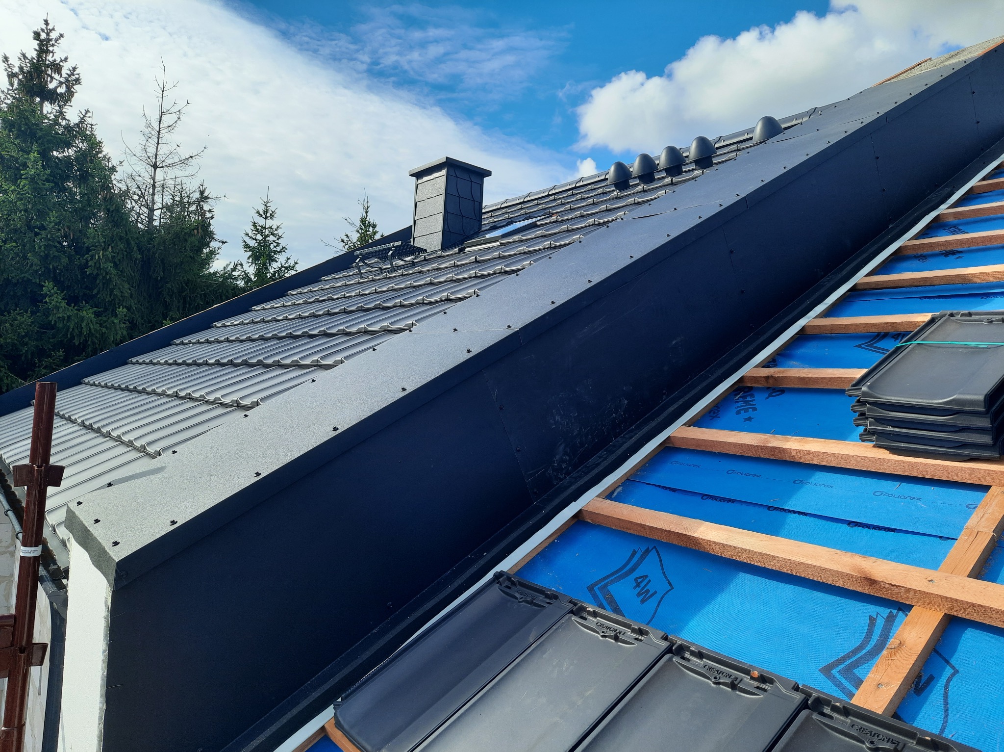 Częściowo pokryty dach z czarną dachówką Creaton, widoczne drewniane łaty i niebieska membrana dachowa, w tle drzewa i błękitne niebo.