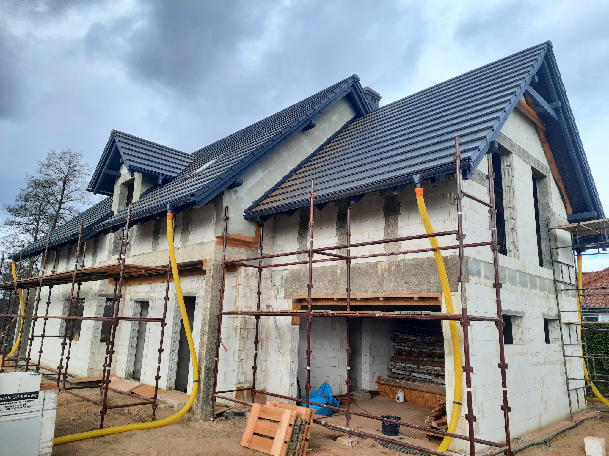 Murowany dom w trakcie budowy z widocznym rusztowaniem, żółtymi rurami wentylacyjnymi, oknami bez szyb i ułożonym ciemnoszarym dachem z dachówki ceramicznej.