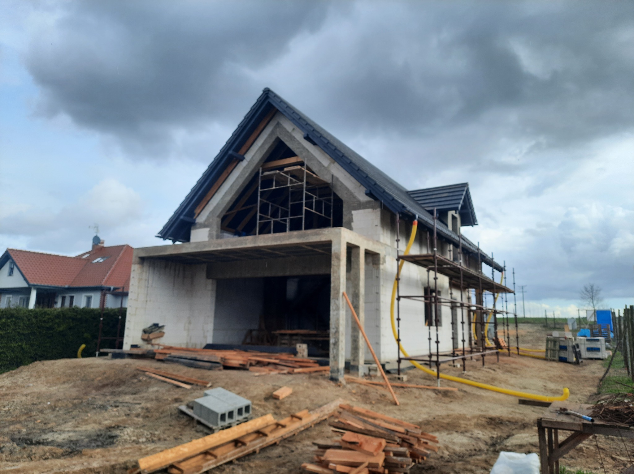 Surowy stan domu z białego pustaka, z czarnym dachem i rusztowaniami, w trakcie budowy, z widocznymi elementami konstrukcyjnymi i stertą drewna na placu budowy.