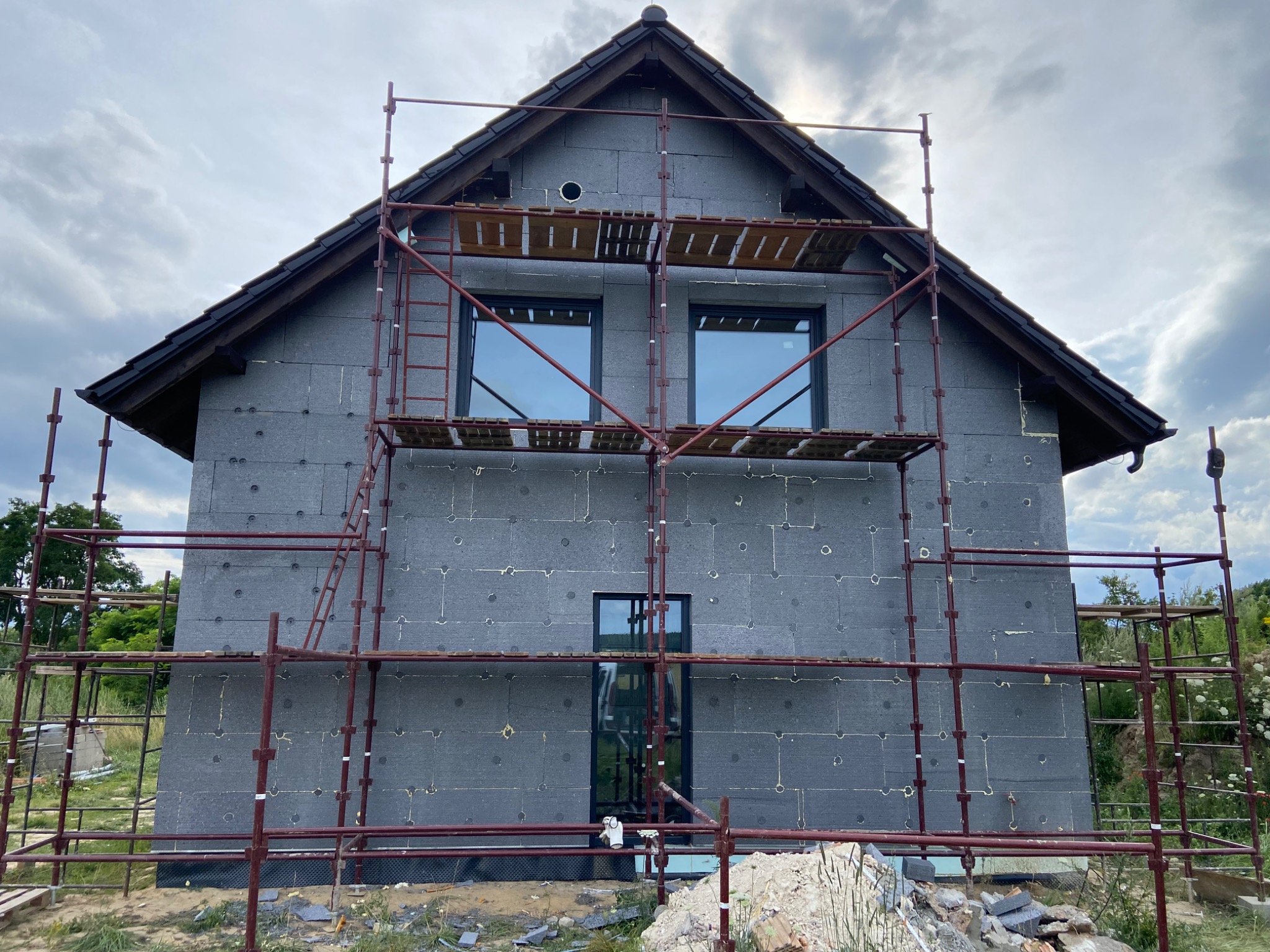 Dwukondygnacyjny dom w trakcie ocieplania styropianem, widoczne rusztowania, okna i dach pokryty dachówką.