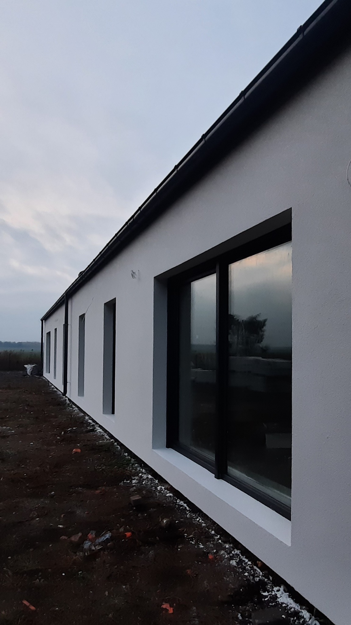 Świeżo otynkowany, parterowy budynek jednorodzinny z czarną stolarką okienną i orynnowaniem, widoczny z niskiej perspektywy, z niezagospodarowanym terenem wokół.