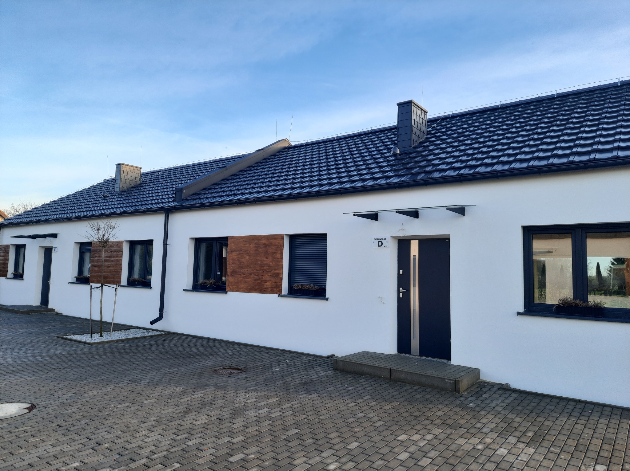 Szeregowy budynek mieszkalny z białymi ścianami, ciemną dachówką i stolarką okienną, wykończony elementami drewnianymi i z kostki brukowej na podjeździe.