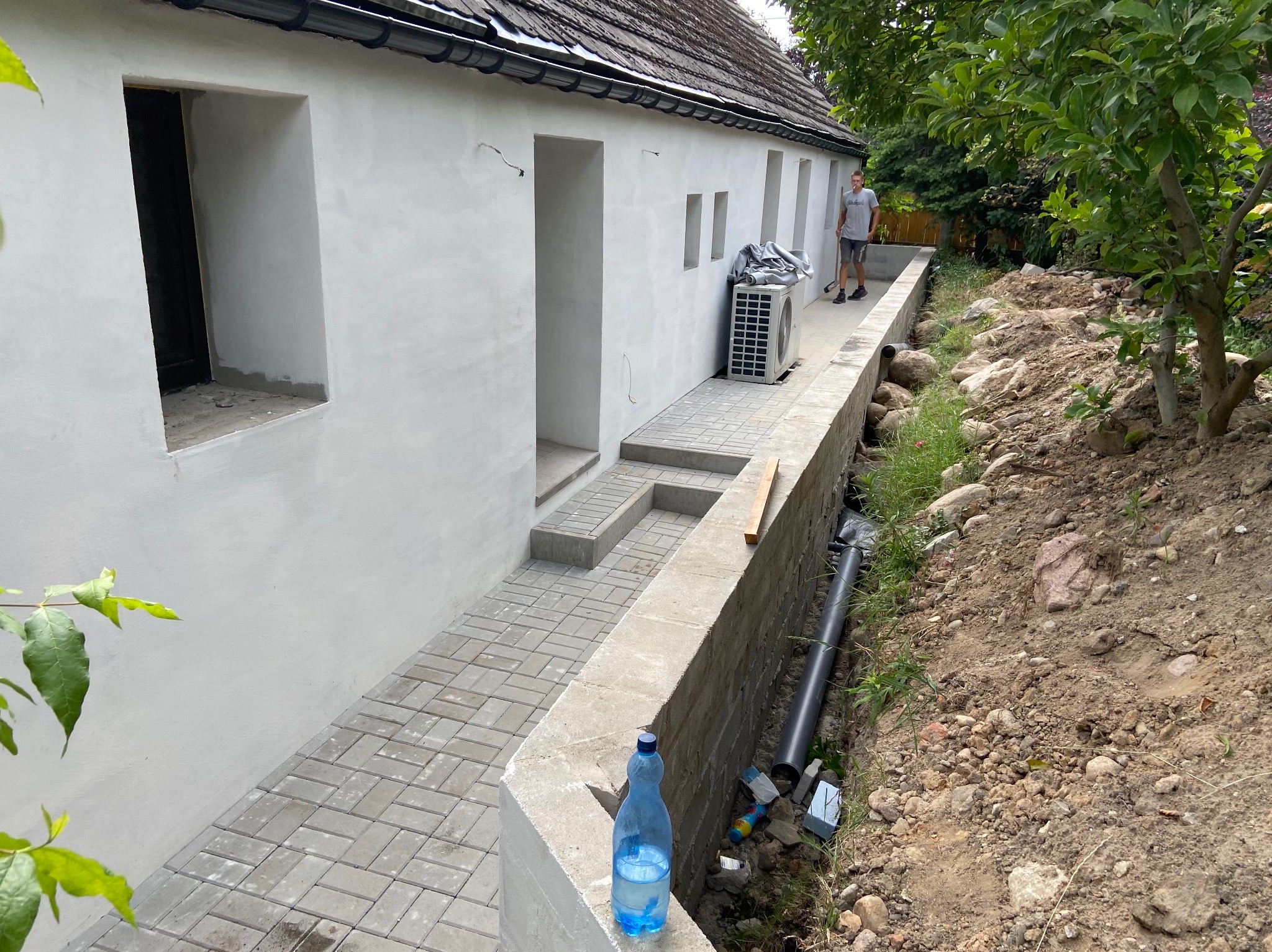 Biała ściana budynku z szarymi elementami chodnika i schodów, widoczna instalacja klimatyzacji i fragment terenu z rurą, w tle mężczyzna z narzędziem.