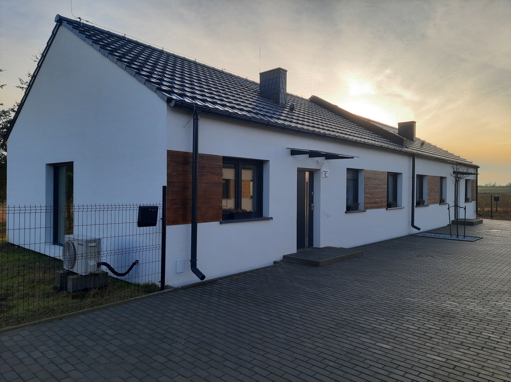 Nowoczesny, parterowy dom szeregowy z białymi ścianami, ciemną dachówką i drewnianymi akcentami na elewacji, widziany z perspektywy chodnika z kostki brukowej o zachodzie słońca.