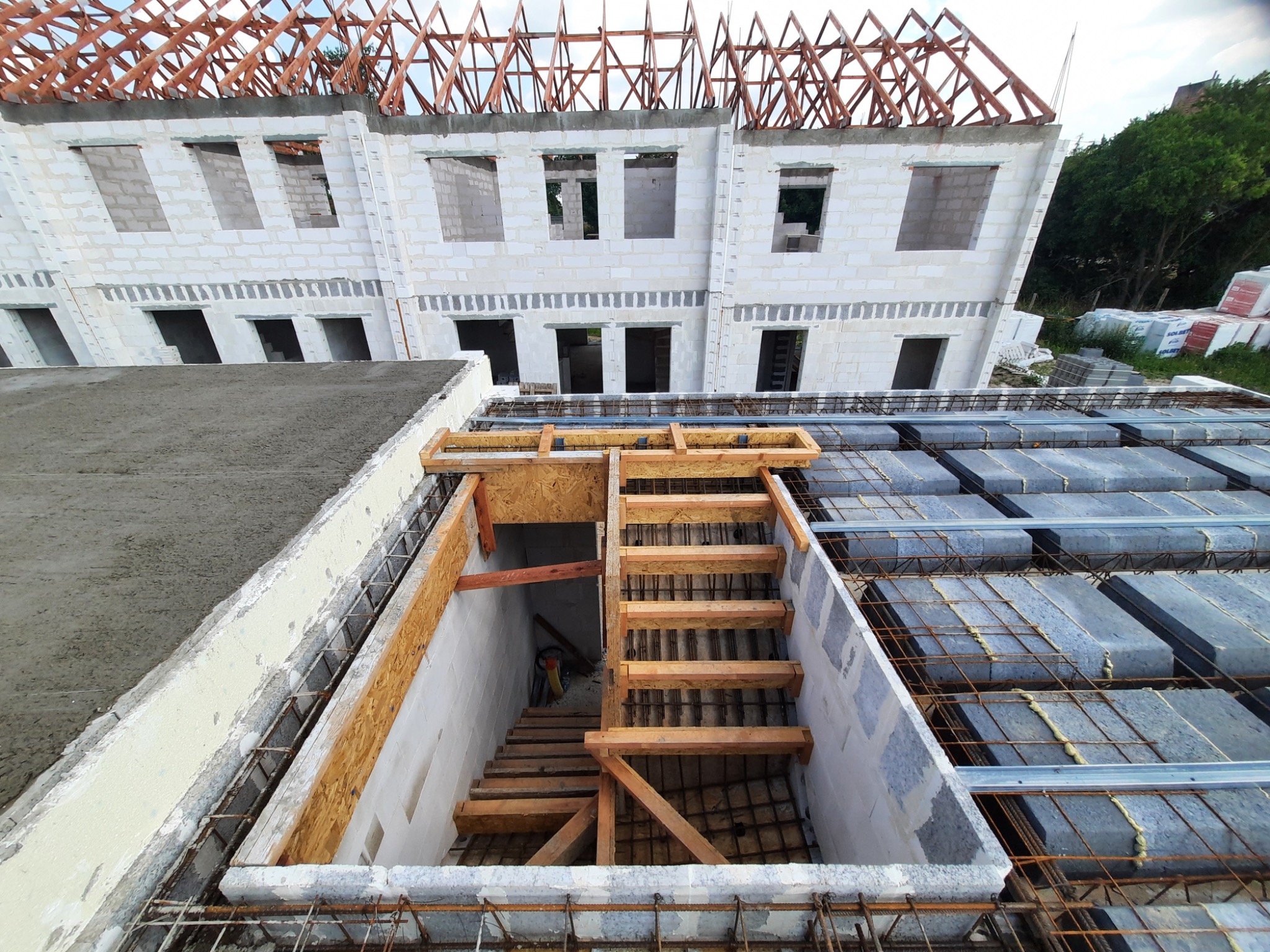 Budowa domu w stanie surowym otwartym: widoczne ściany z betonu komórkowego, więźba dachowa, betonowy strop z deskowaniem i schody prowadzące do piwnicy.