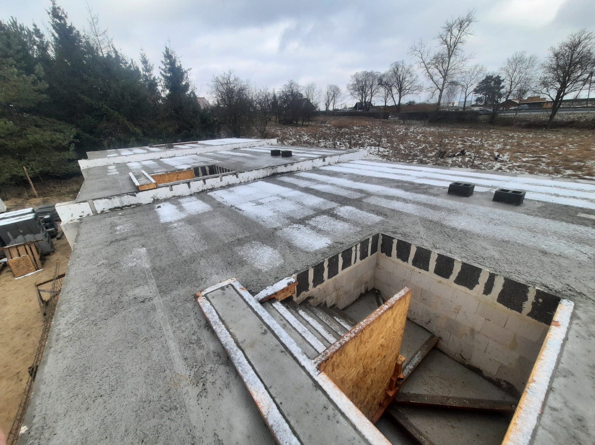 Wylany betonowy strop z widocznym otworem na schody, zabezpieczonym płytą OSB, lekko przyprószony śniegiem, w tle drzewa i pochmurne niebo.