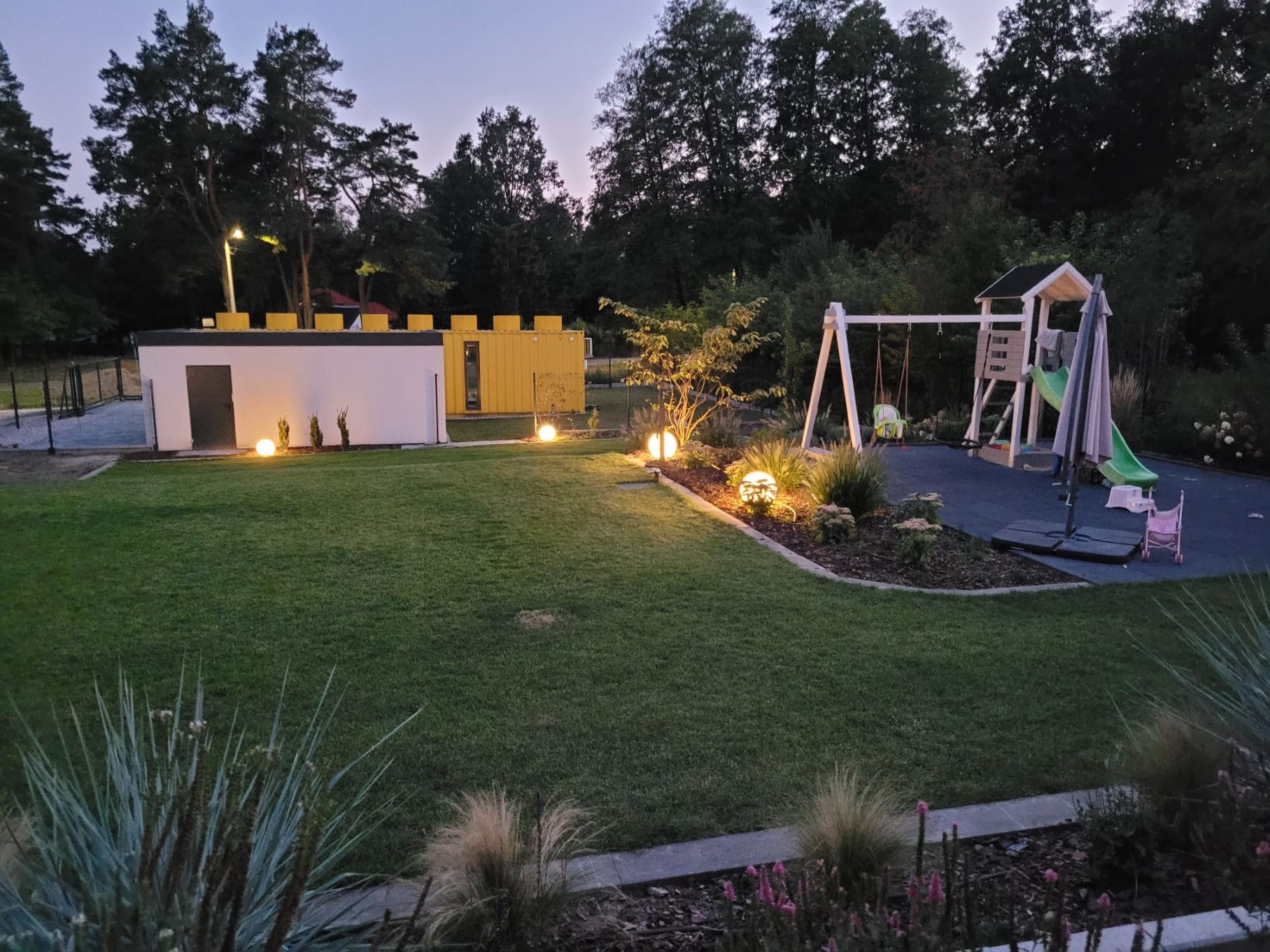 Ogród z równo skoszoną trawą o zmierzchu, oświetlony kulistymi lampami, z nowoczesnym domkiem w tle i placem zabaw z huśtawkami i zjeżdżalnią.