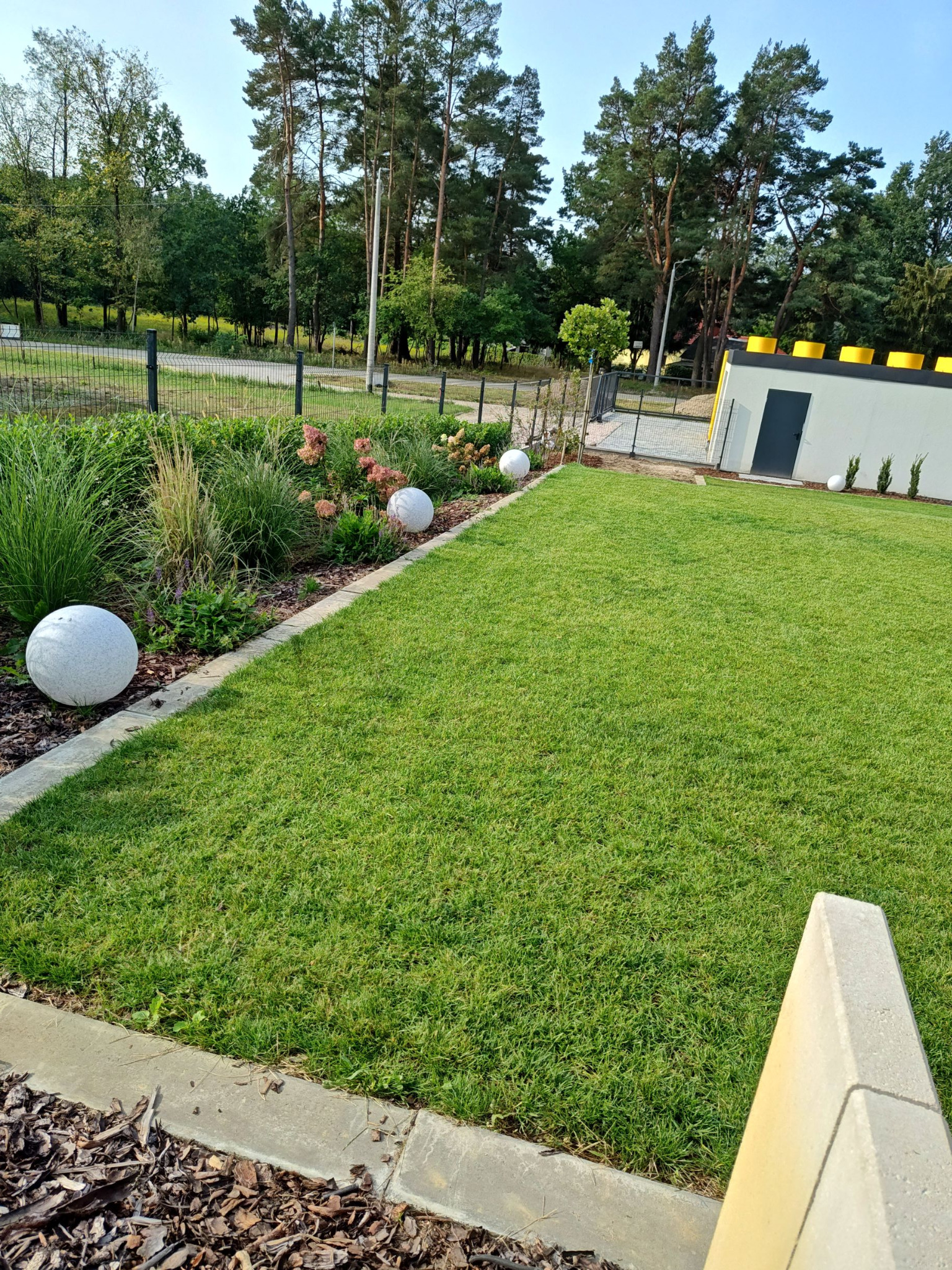 Starannie utrzymany trawnik z dekoracyjnymi kulami i rabatą kwiatową, widok na dom z elementami przypominającymi klocki Lego.