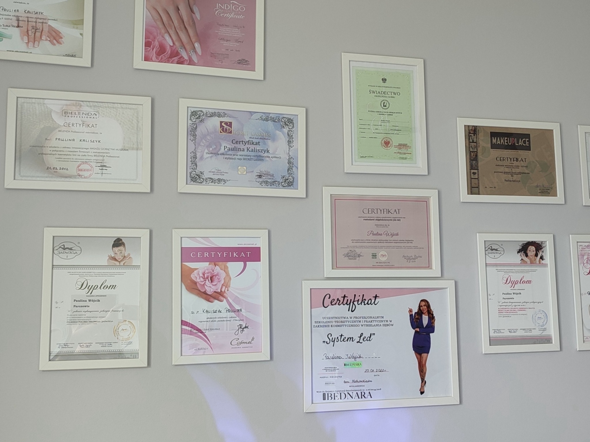 Ściana w salonie kosmetycznym z ramkami zawierającymi dyplomy i certyfikaty ukończonych szkoleń i kursów kosmetycznych, w tym certyfikaty z zakresu stylizacji paznokci, przedłużania rzęs...