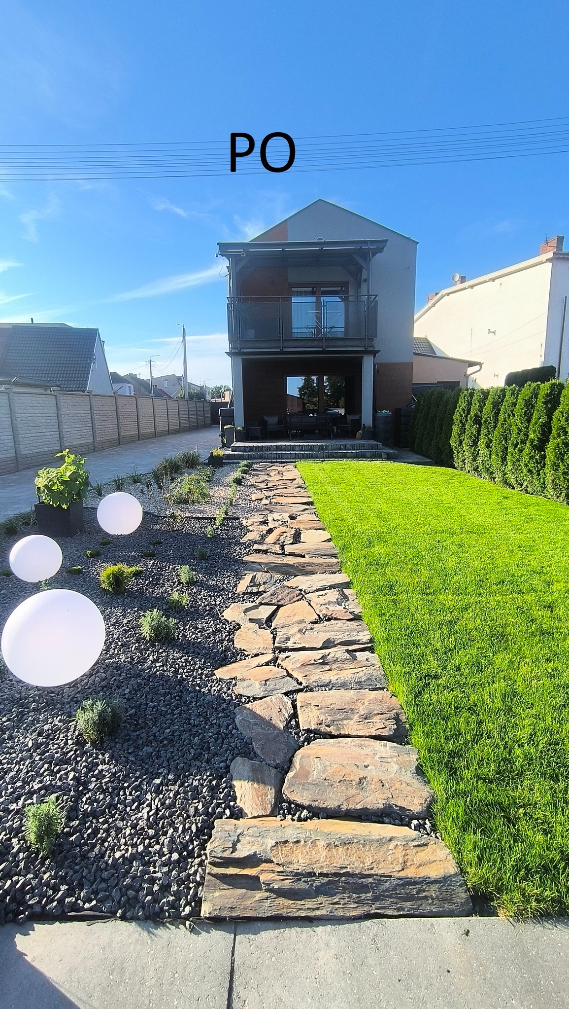 Ogród po realizacji: ścieżka z kamienia prowadzi przez trawnik, obok rabata z kamykami i kulistymi lampami. W tle nowoczesny dom z balkonem.