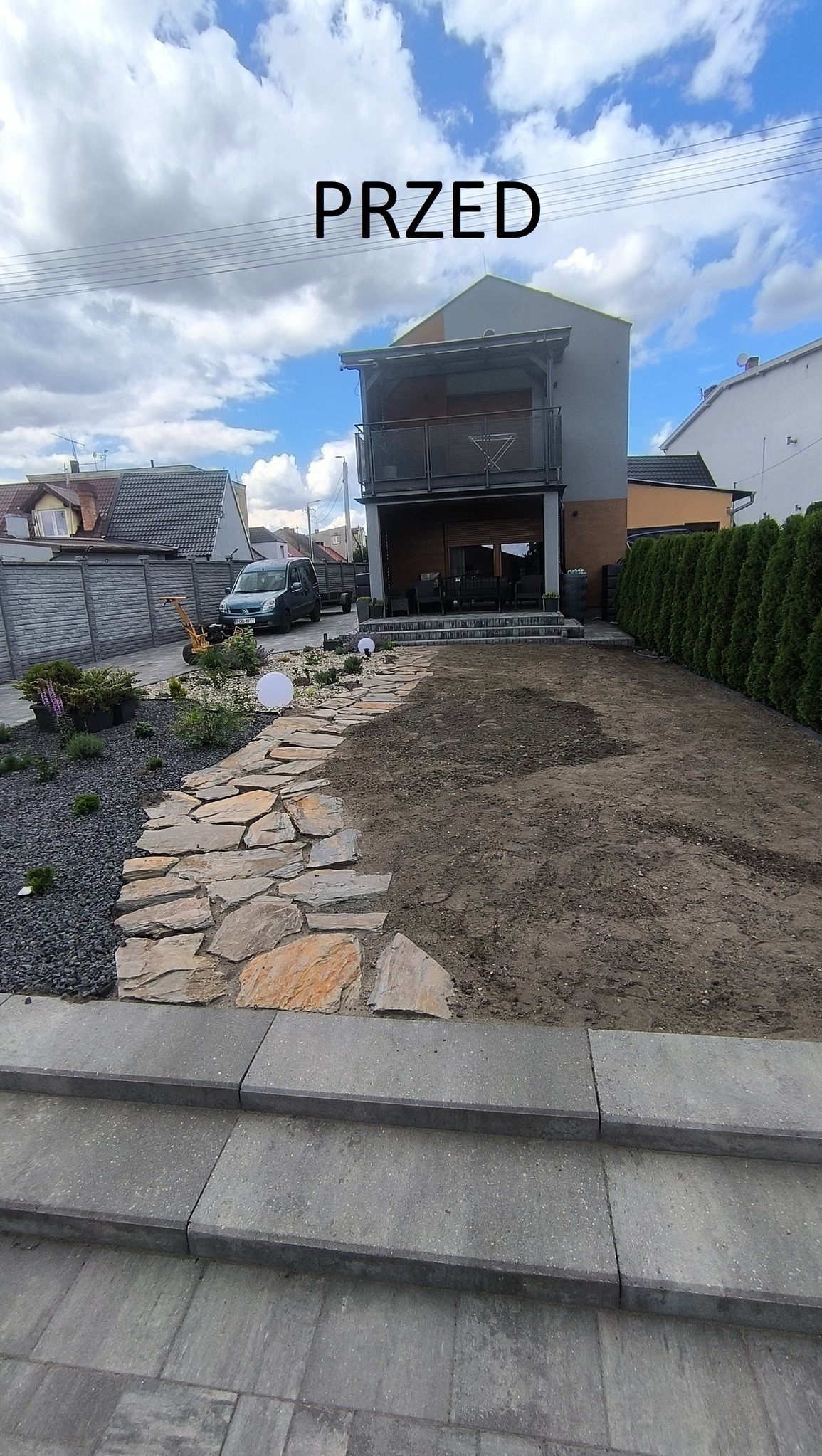Etap przygotowania terenu pod ogród z wytyczoną ścieżką z kamienia łupanego, widok na dom z balkonem i fragment żywopłotu.